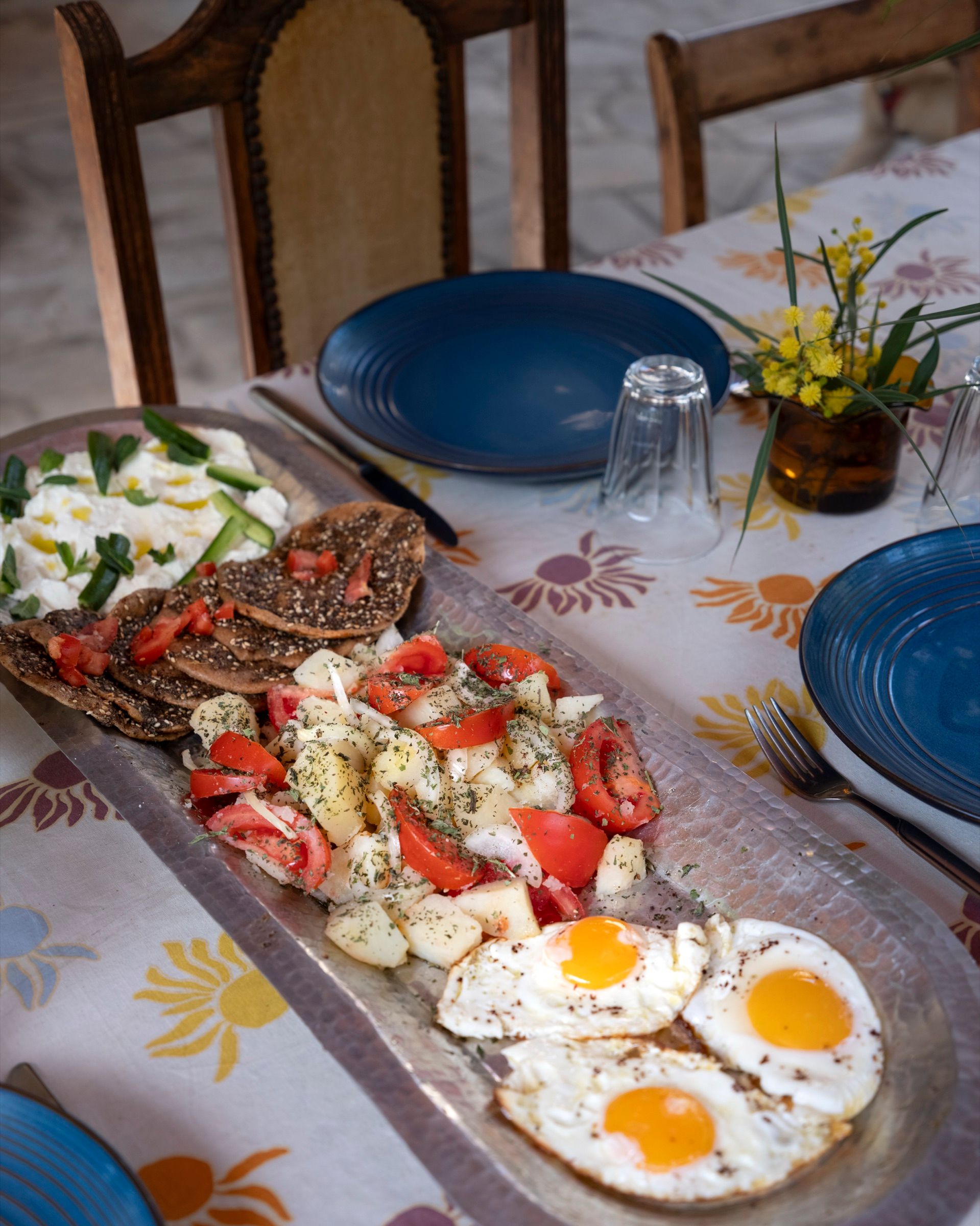 A breakfast table laid with fried eggs, tomato and cucumber salad, bread and blue ceramic bowls
