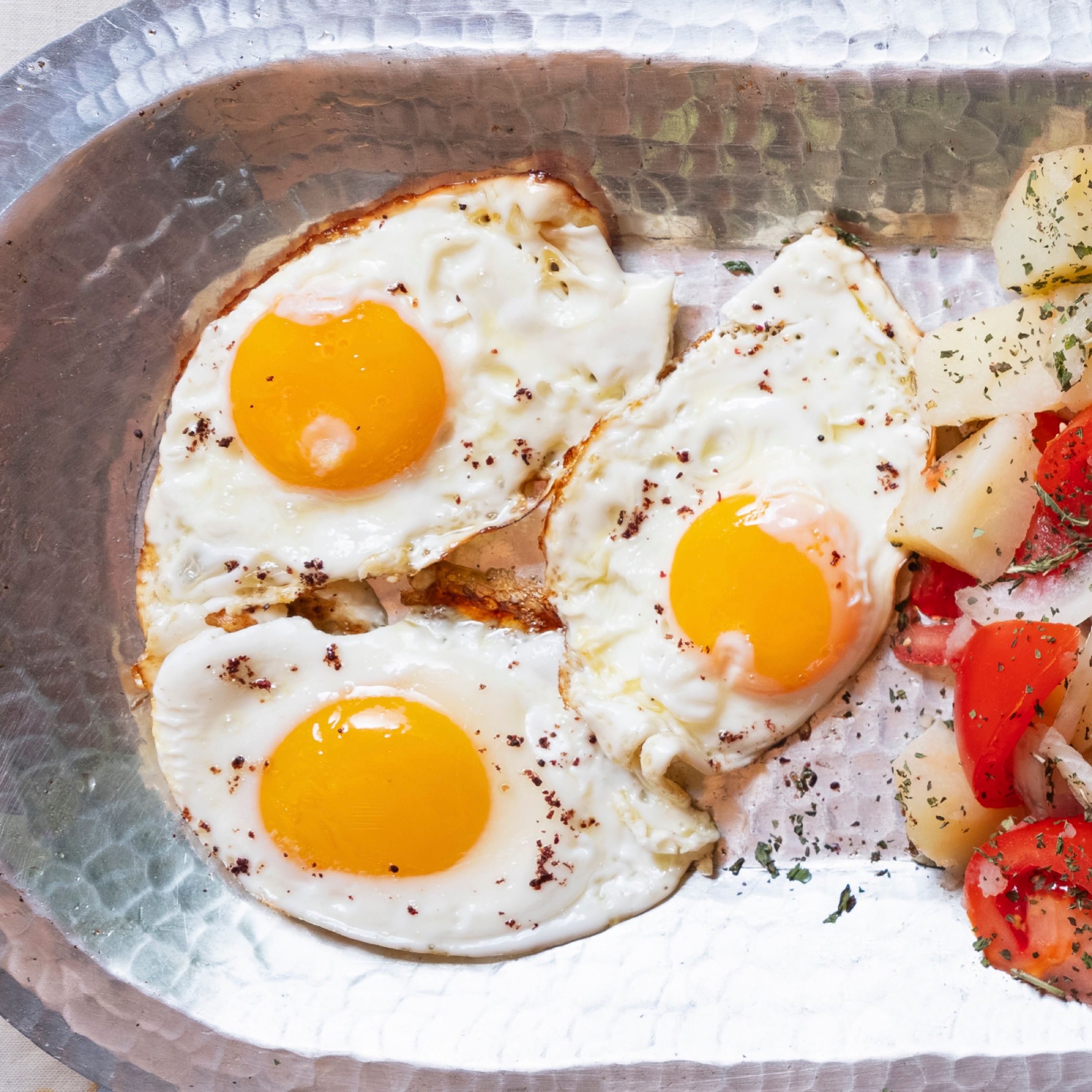 Three fried eggs dusted with sumac on a hammered copper tray, served with potatoes, tomato and herbs