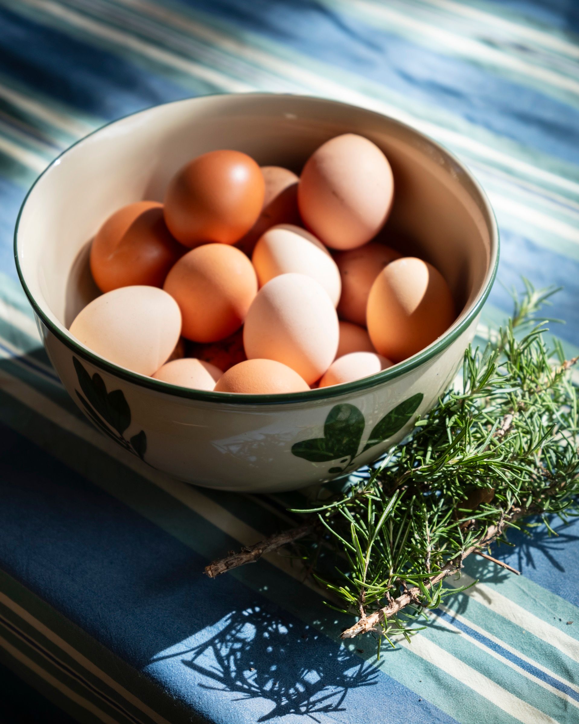 A ceramic bowl of brown eggs beside a bunch of fresh thyme on a striped cloth