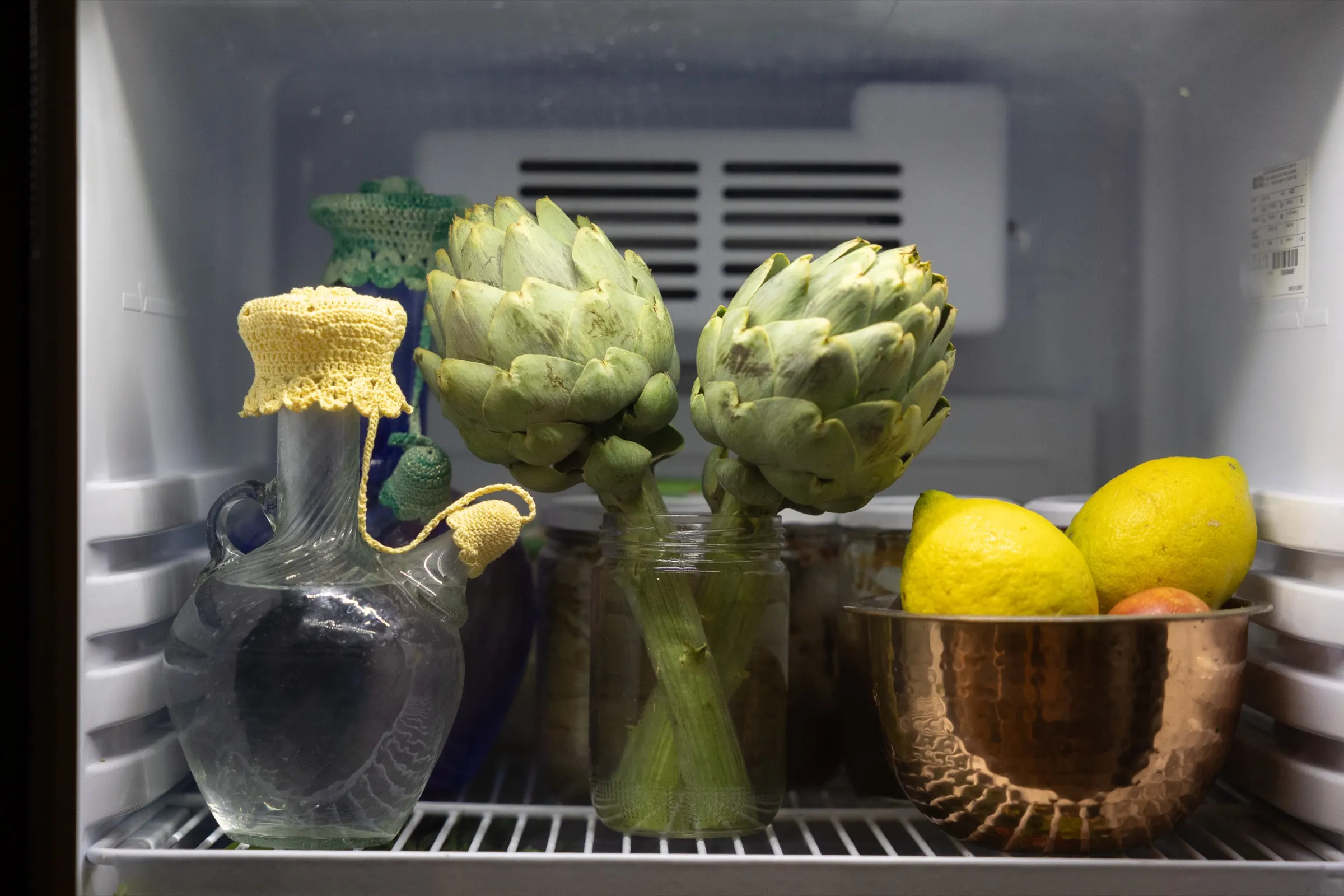 An open refrigerator shelf filled with garden produce — artichokes, lemons in a woven bowl, herbs and greens