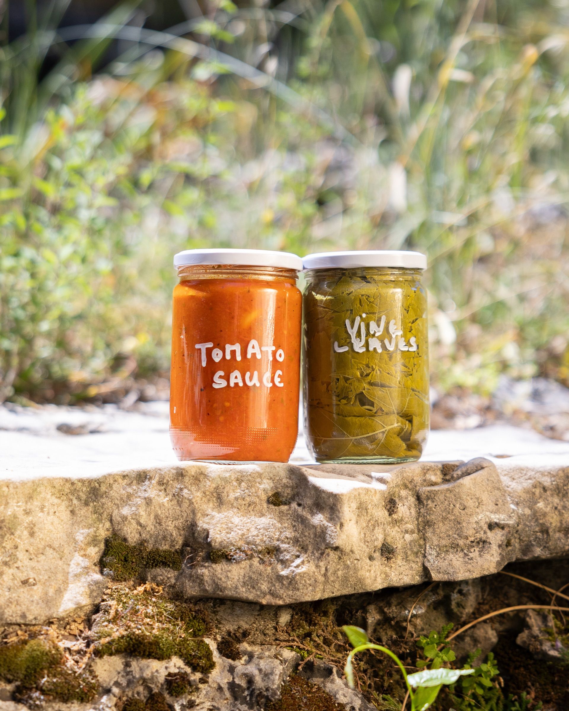 Two jars of homemade preserves labelled by hand — tomato sauce and vine leaves — standing on a stone wall