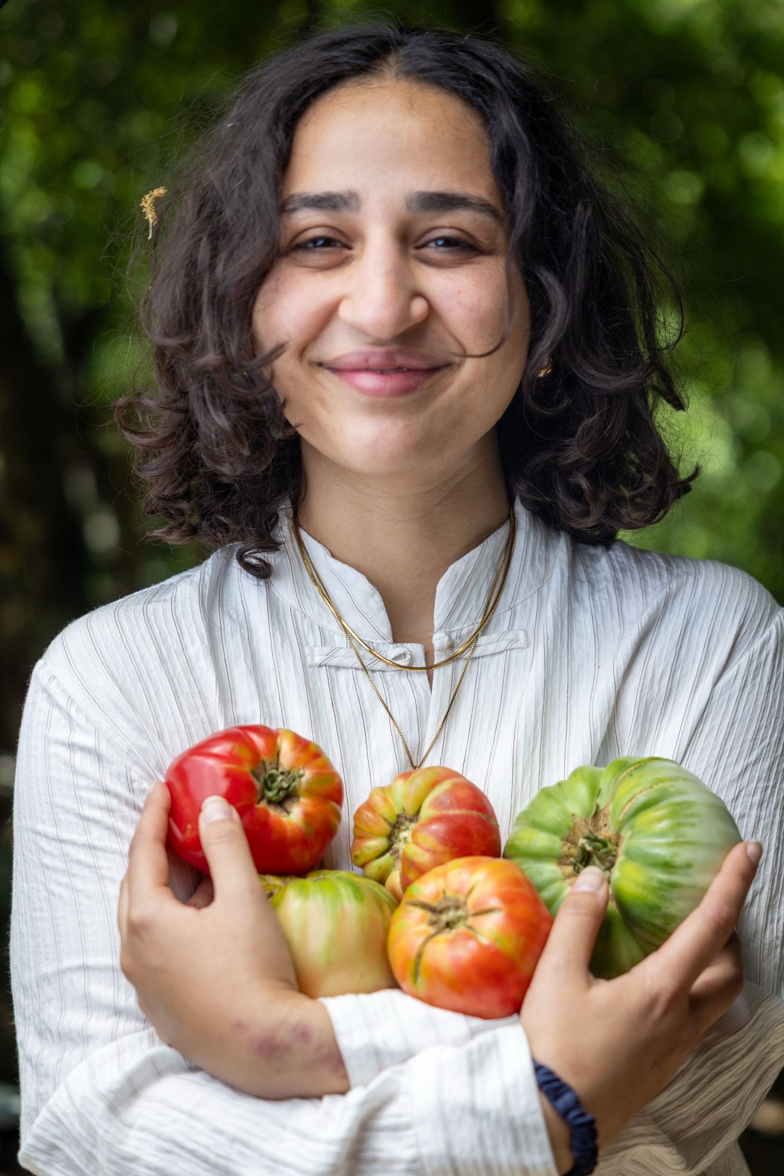 A woman in a white shirt smiling as she holds an armful of heirloom tomatoes just picked from the garden