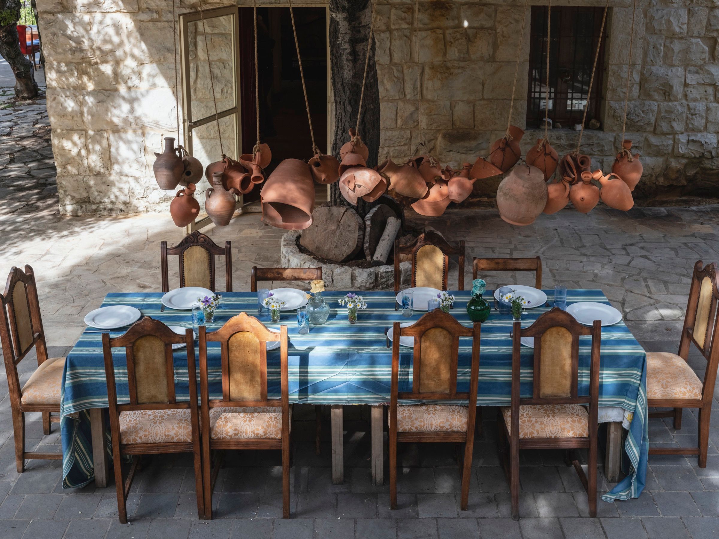 A long outdoor table dressed in blue-striped cloth and set with earthenware, beside a wall of stacked terracotta jars