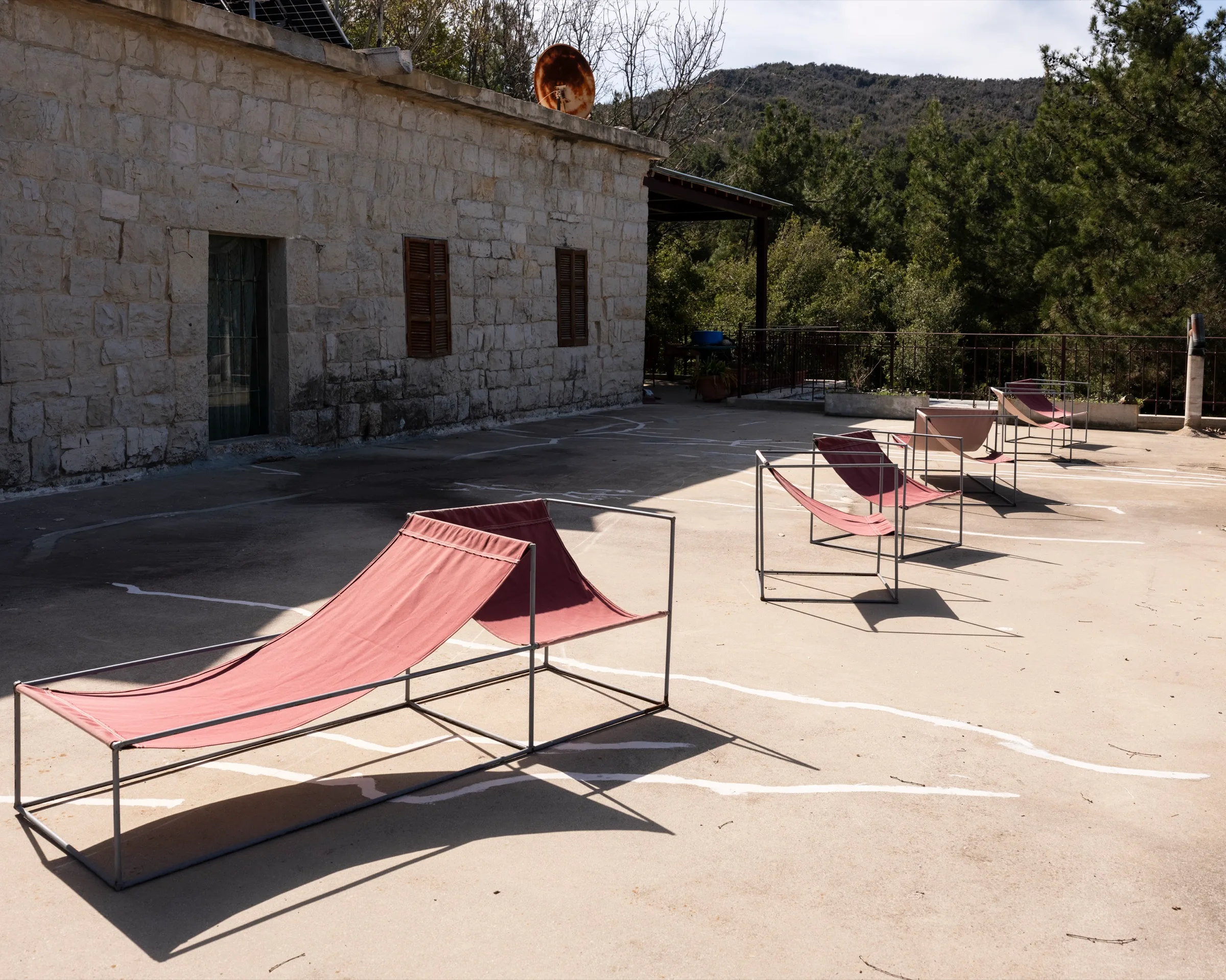 The stone façade of the guesthouse seen across its terrace, with canvas sling chairs in the foreground and pine-covered mountains rising behind