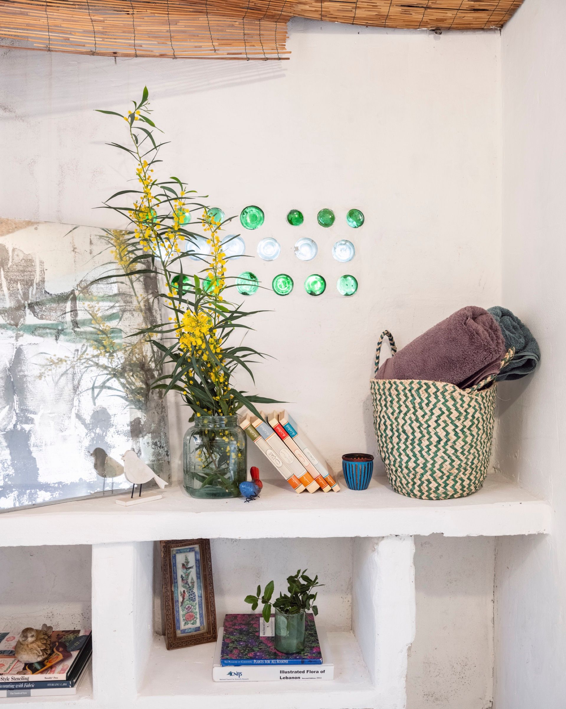 A whitewashed alcove in the Thyme room, with yellow mimosa in a glass jar, folded towels in a woven basket, and green and clear bottle bases set into the wall