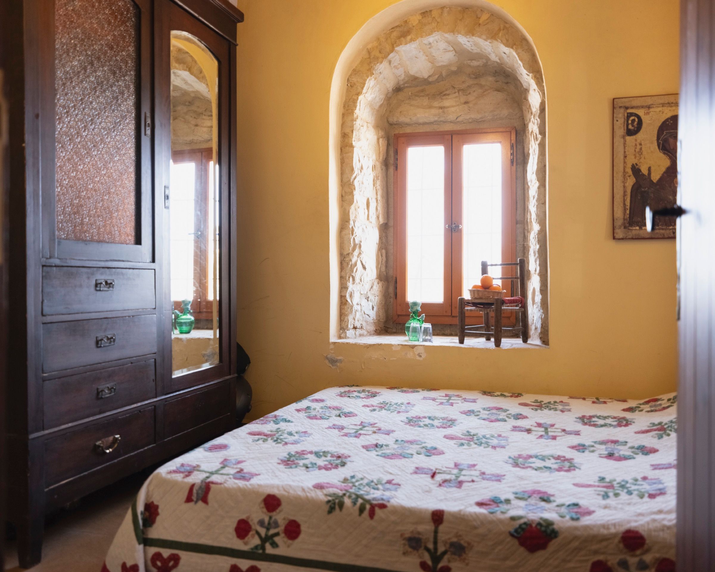 A bedroom with deep ochre walls, an arched stone window recess and an antique wooden wardrobe, the bed covered in an embroidered floral quilt
